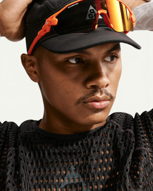 Nike ACG men's black open-hole mesh Dri-FIT race-day top worn by a model in a black cap and orange sports sunglasses.
