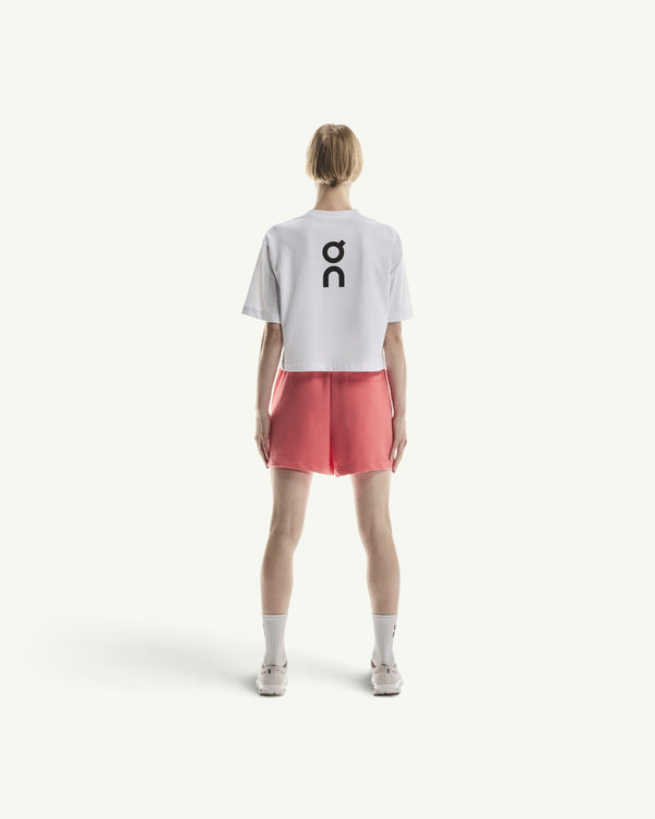 On women's white boxy cotton-blend t-shirt with black logo on back, paired with coral shorts, rear view