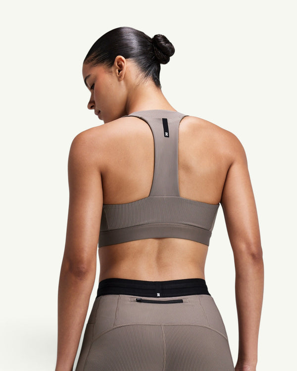 Amateur women's taupe ribbed T-back sports bra with racerback design and branded detail, worn by a model from behind.