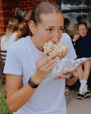 Amateur Women's Twilight Tee in light blue, a silky-soft technical knit shirt worn by a woman eating a bagel.